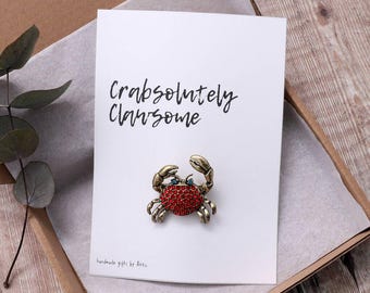 May include: A white card with the text "Crabsolutely Clawesome" and a red jeweled crab pin.