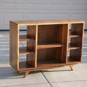 May include: A wooden entertainment center with a mid-century modern design. It has a dark walnut finish and features multiple shelves for displaying items. The unit has four legs that are angled outward.