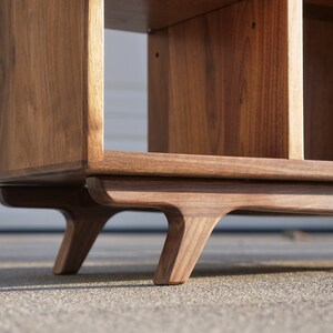 May include: A wooden shelf unit with a mid-century modern design. The shelf has a rectangular shape with open shelves and a solid back. The shelf is supported by four angled legs.