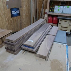 May include: A collection of unfinished hardwood boards in varying sizes, stacked on a workbench. The wood has a natural grain and a light brown color. Some boards have blue tape on the edges. The scene is set in a workshop.