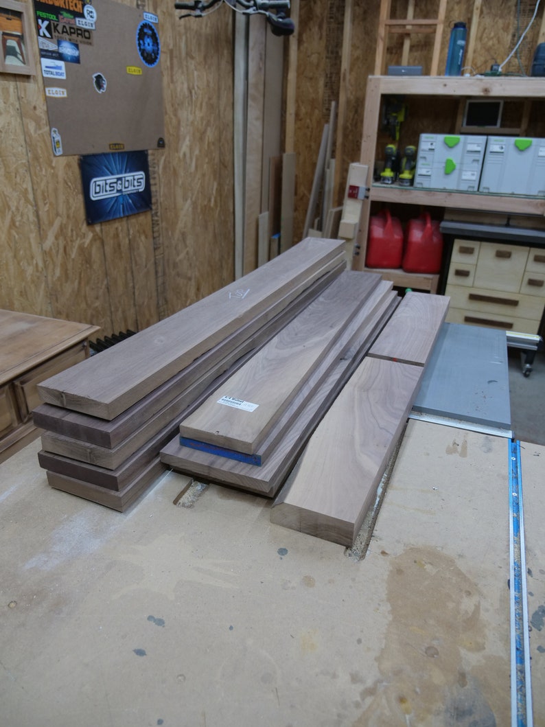 May include: A collection of unfinished hardwood boards in varying sizes, stacked on a workbench. The wood has a natural grain and a light brown color. Some boards have blue tape on the edges. The scene is set in a workshop.