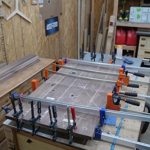 May include: A woodworking project in progress, featuring a large, dark wood panel clamped with multiple bar clamps. The panel has white markings and drilled holes. The workshop setting includes tools, lumber, and a workbench.