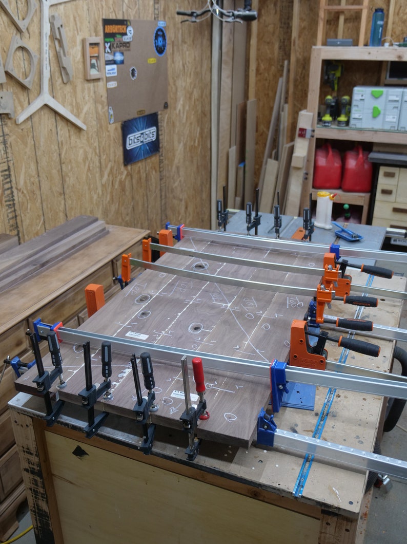 May include: A woodworking project in progress, featuring a large, dark wood panel clamped with multiple bar clamps. The panel has white markings and drilled holes. The workshop setting includes tools, lumber, and a workbench.