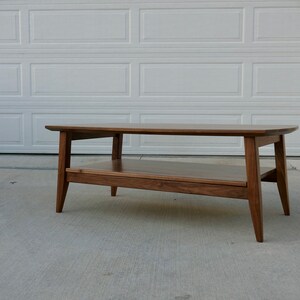 May include: A mid-century modern style coffee table made of dark wood. The table has a rectangular top with rounded corners, a lower shelf, and angled legs. The table measures approximately 48 inches long, 20 inches wide, and 16 inches high.