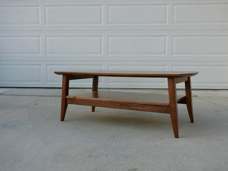 May include: A mid-century modern style coffee table made of dark wood. The table has a rectangular top with rounded corners, a lower shelf, and angled legs. The table measures approximately 48 inches long, 20 inches wide, and 16 inches high.