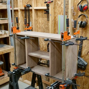 May include: A wooden cabinet with two shelves is being assembled using clamps and sawhorses. The cabinet is made of walnut wood and has a natural finish. The cabinet is being assembled in a workshop with tools and other woodworking supplies visible in the background.