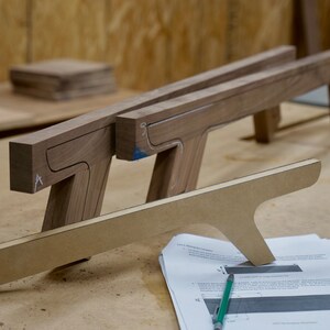 May include: Two wooden pieces with cutouts, likely for a furniture project, are laid out on a workbench. A sheet of paper with instructions and a green pencil are nearby.