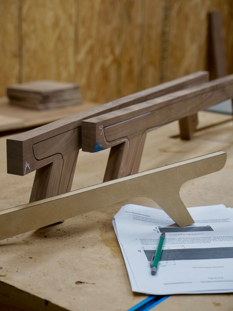 May include: Two wooden pieces with cutouts, likely for a furniture project, are laid out on a workbench. A sheet of paper with instructions and a green pencil are nearby.