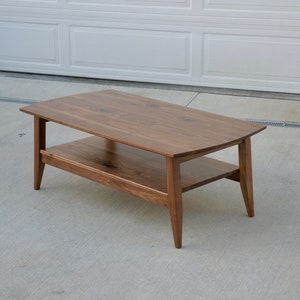 May include: A rectangular coffee table made of dark brown wood. The table has a lower shelf and angled legs. The table is approximately 48 inches long, 24 inches wide, and 18 inches high.