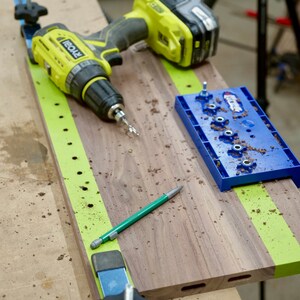 May include: A green and yellow drill, a blue jig, and a green pencil on a wooden workbench. The jig has a series of holes for drilling.
