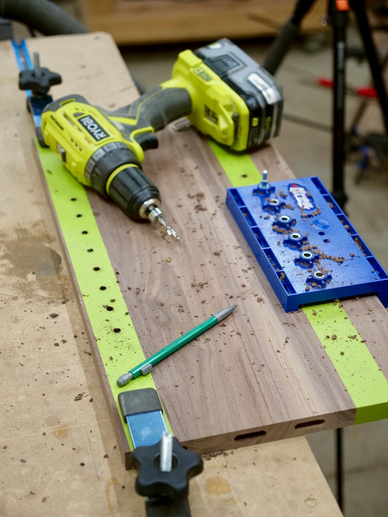 May include: A green and yellow drill, a blue jig, and a green pencil on a wooden workbench. The jig has a series of holes for drilling.