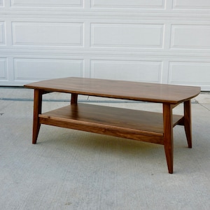 May include: A rectangular, mid-century modern coffee table made of dark wood. The table has a lower shelf and angled legs. The table is approximately 48 inches long, 20 inches wide, and 16 inches high.
