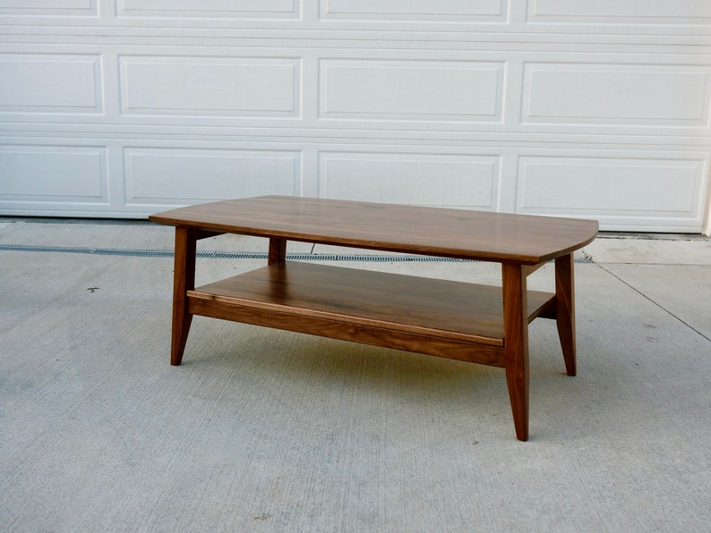 May include: A rectangular, mid-century modern coffee table made of dark wood. The table has a lower shelf and angled legs. The table is approximately 48 inches long, 20 inches wide, and 16 inches high.