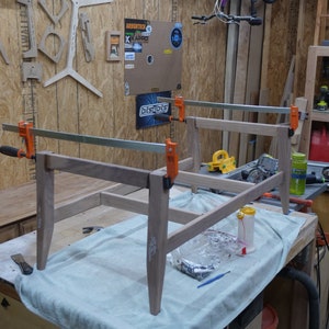 May include: A partially assembled wooden table frame clamped together with orange clamps. The light-colored wood is on a workbench covered with a light blue cloth. Tools and materials are nearby, suggesting a woodworking project.