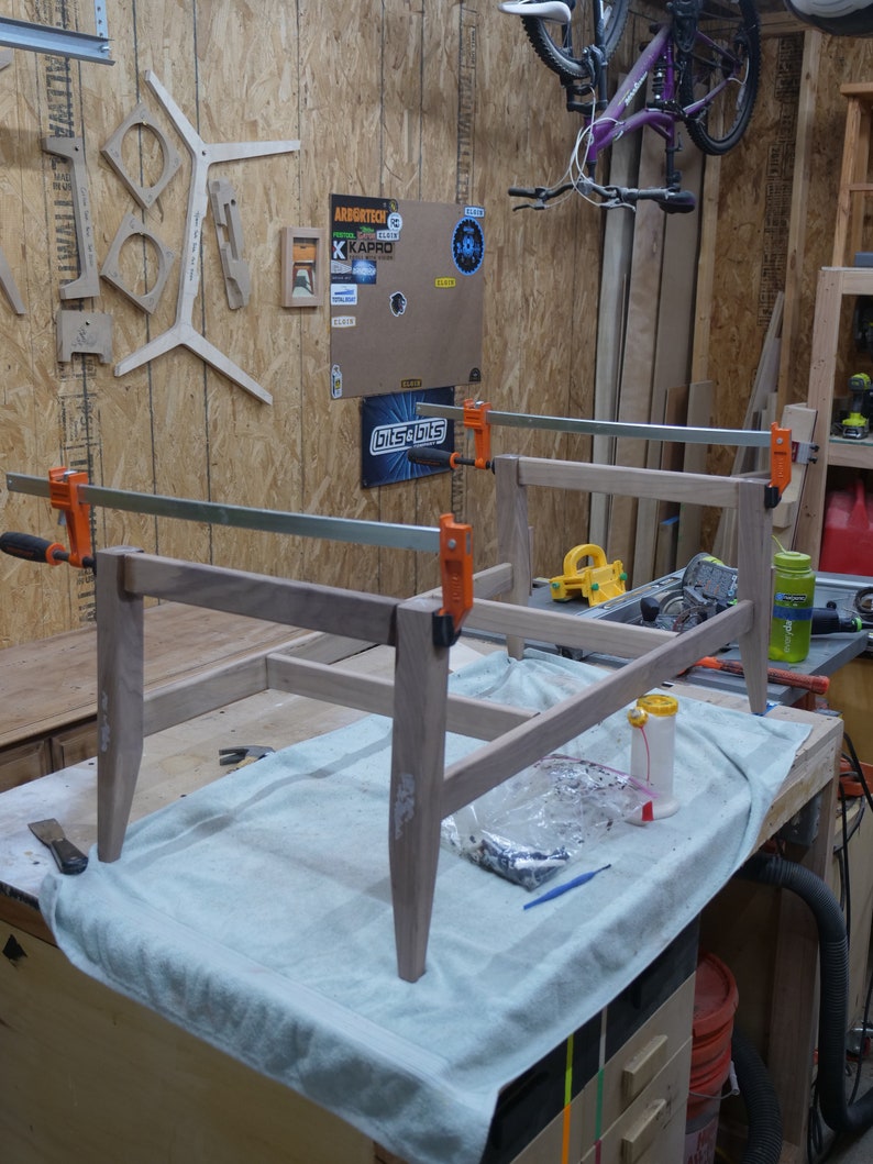 May include: A partially assembled wooden table frame clamped together with orange clamps. The light-colored wood is on a workbench covered with a light blue cloth. Tools and materials are nearby, suggesting a woodworking project.