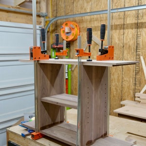 May include: A wooden shelf unit with two shelves is being assembled using orange and black clamps. The shelf unit is made of walnut wood and has a natural finish.