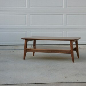 May include: A mid-century modern style coffee table made of wood. The table has a rectangular top with rounded corners, a lower shelf, and angled legs. The wood has a warm, brown tone.