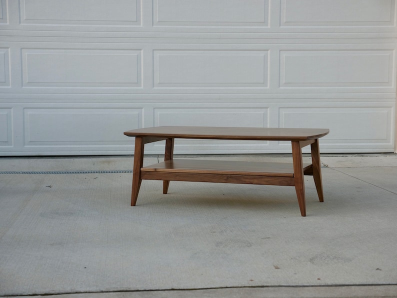 May include: A mid-century modern style coffee table made of wood. The table has a rectangular top with rounded corners, a lower shelf, and angled legs. The wood has a warm, brown tone.