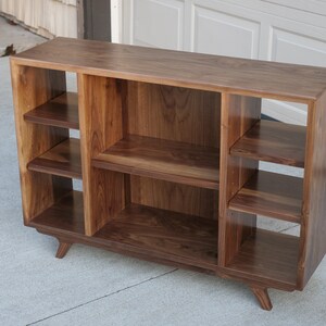 May include: A walnut wood record player stand with three open shelves on each side and a lower shelf. The stand has a mid-century modern design with angled legs.