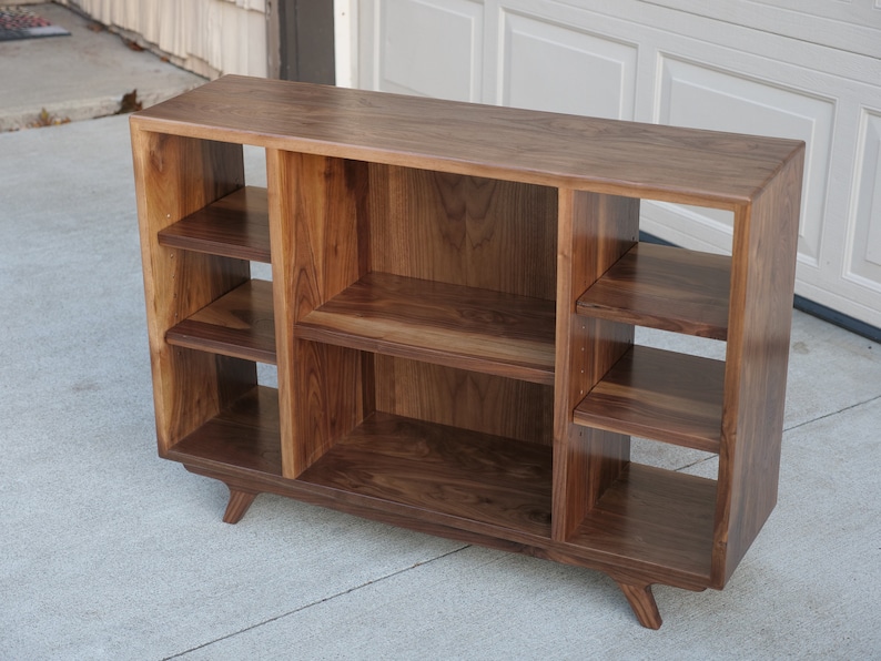 May include: A walnut wood record player stand with three open shelves on each side and a lower shelf. The stand has a mid-century modern design with angled legs.