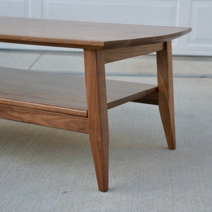 May include: A mid-century modern style coffee table made of dark brown wood. The table has a rounded top and a lower shelf for storage. The legs are tapered, and the table is set on a concrete surface.