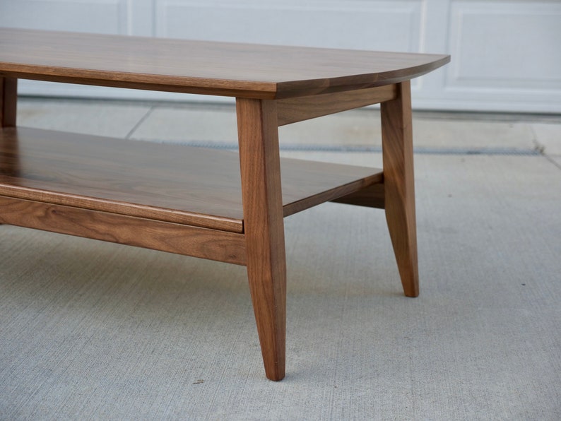 May include: A mid-century modern style coffee table made of dark brown wood. The table has a rounded top and a lower shelf for storage. The legs are tapered, and the table is set on a concrete surface.