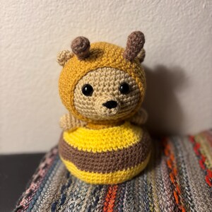 May include: A handmade crochet bee plush toy. The toy features a tan body with a yellow and brown striped bottom. It wears a yellow hood with brown antennae and has black eyes and a black nose.