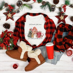 May include: White t-shirt with a Christmas-themed design featuring a gingerbread house, a Christmas tree, a mug of hot chocolate, and the text "Hot Chocolate With Cookies And Watching Christmas Movies". The shirt is displayed with a red and black plaid shirt, a pair of brown suede boots with white fur trim, a red and black plaid scarf, a pair of blue jeans, and a red to-go cup.