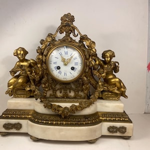 May include: An antique gold-toned clock with a white face and Roman numerals. Two gold cherub figures flank the clock face, and the clock sits on a white and gold marble base. The clock features ornate detailing and a decorative floral garland.