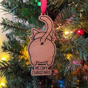May include: A wooden Christmas ornament shaped like a cat's rear end with a tail curled over its back. The ornament is brown and has a black outline. The text "MEOWY CHRISTMAS" is printed on the bottom of the ornament.