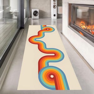May include: A rectangular rug with a cream background and a retro-style design. The design features a wavy, colorful pattern in shades of red, orange, yellow, blue, and green. The rug is placed on a polished concrete floor.