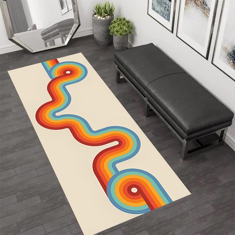 May include: A rectangular rug with a cream background and a retro design. The design features a wavy, colorful pattern of concentric rainbow-colored lines in red, orange, yellow, blue, and green. The rug is placed on a dark wood floor.