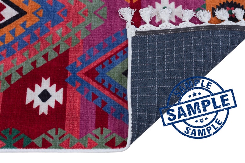 May include: A colorful rug with a geometric pattern in shades of red, blue, green, and orange. The rug has a white fringe border and a gray backing. The word "SAMPLE" is stamped in blue on the backing.