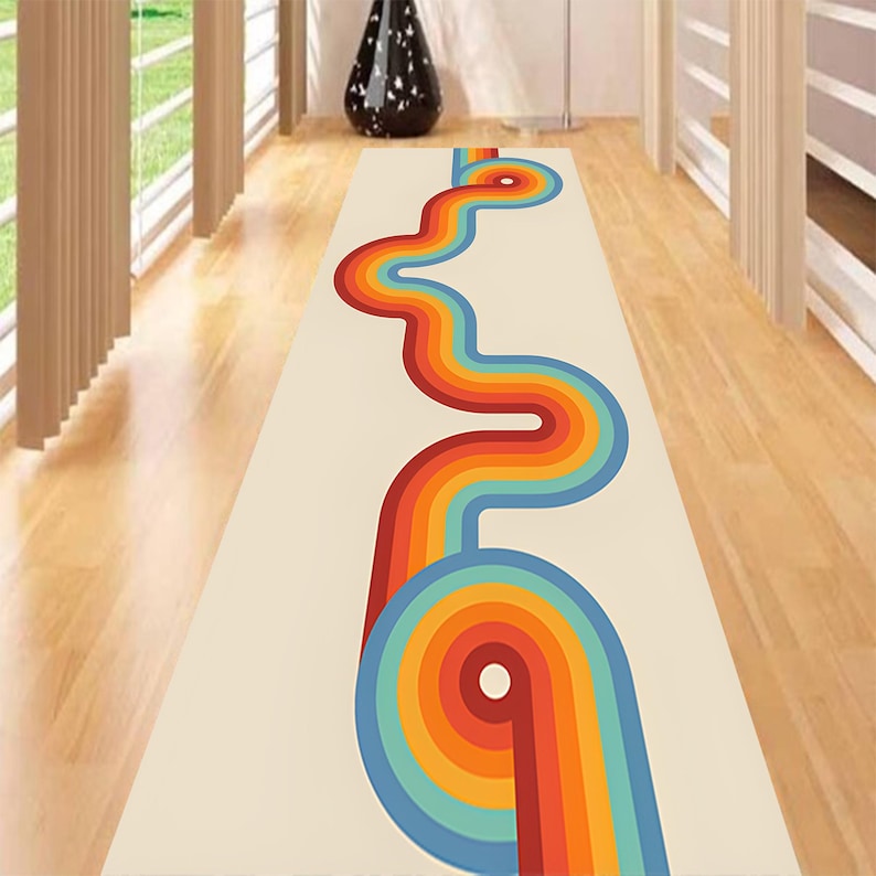 May include: A rectangular cream-colored rug with a retro design. A wavy, colorful pattern in shades of red, orange, yellow, and blue runs down the center. The rug is placed on a wooden floor.