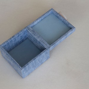 May include: A light blue, two-compartment box with a crocodile-embossed pattern. The box is open, revealing a blue interior.