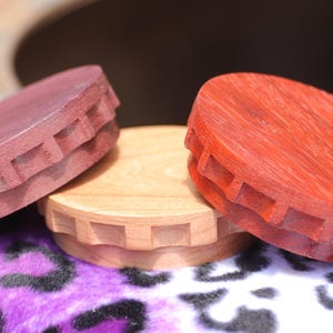May include: Three wooden bottle stoppers in shades of red, purple, and natural wood. Each stopper has a circular top with a decorative, notched edge. The stoppers are displayed on a purple and black patterned surface.