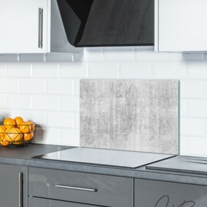 May include: A gray concrete-look glass splashback for a kitchen stovetop. The splashback is mounted above a white countertop and a gray cabinet with silver handles.