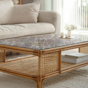 Puede incluir: Mesa de centro rectangular con tapa de cristal con un diseño estampado en tonos azules, rojos y beige. La mesa tiene un marco de ratán natural y un estante inferior. Velas y un jarrón con flores encima.