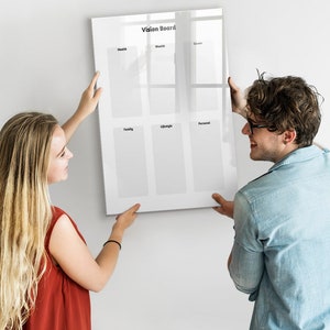 Op de afbeelding: Twee mensen hangen een wit vision board aan een muur. Het bord is verdeeld in zes secties met de koppen: Gezondheid, Welvaart, Carrière, Familie, Lifestyle en Persoonlijk.