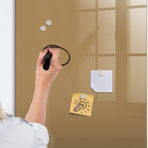 Op de afbeelding: Een persoon schrijft op een gouden magnetisch whiteboard met een zwarte marker. Er zijn twee witte magneten en twee sticky notes op het bord. Een sticky note heeft een tekening van een gloeilamp.