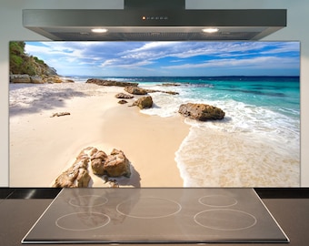 Puede incluir: Un protector de salpicaduras de cocina con una imagen panorámica de una playa de arena con agua turquesa y rocas. La imagen está detrás de una encimera de cocina negra con cuatro quemadores.