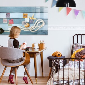 Könnte beinhalten: Ein Kind sitzt an einem Schreibtisch mit einem Holzstuhl und einer Lampe. An der Wand hängt eine blaue und weiße Holztafel mit einem Seil, Muscheln und einem Seestern. Die Tafel ist mit bunten Haftnotizen mit Text versehen. Das Kind hält eine kleine weiße Flasche.