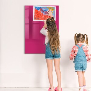Op de afbeelding: Twee kinderen, een met een spijkerbroek overall en een geruit shirt, en de ander met een spijkerrok en een wit shirt, staan voor een roze magnetisch whiteboard. Het bord heeft een kleurrijke tekening erop.