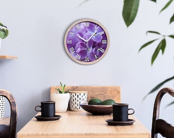 May include: A round wall clock with a purple flower design and Roman numeral markings. The clock is hanging on a light grey wall in a room with a wooden table and chairs.