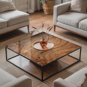 May include: A square coffee table with a dark wood grain top and a black metal frame. A small glass vase with dried plants sits on a wooden tray on the table. The table is in a living room setting with neutral-toned furniture.