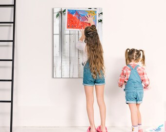 May include: Two girls, one wearing a denim overall dress and the other wearing a denim skirt and a white shirt, are hanging a framed painting on a white wall. The painting is a colorful abstract piece with a white background.