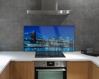 Puede incluir: Un protector contra salpicaduras de cocina con una vista nocturna de la ciudad de Nueva York, con el puente de Brooklyn y rascacielos iluminados. La imagen tiene una combinación de colores azul y blanco, con las luces de la ciudad reflejadas en el agua.