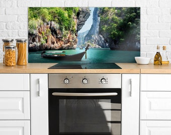 Puede incluir: Un protector de salpicaduras de cocina con una imagen panorámica de un barco de cola larga en una laguna tropical rodeada de acantilados verdes exuberantes. El protector de salpicaduras está instalado sobre un horno de acero inoxidable con perillas negras.