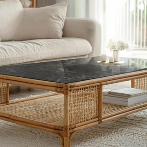 Op de afbeelding: Een rechthoekige salontafel met een zwart marmerlook blad en een natuurlijk rotan frame. De tafel heeft een onderste plank voor opslag en is versierd met kaarsen en een vaas met bloemen. De tafel is een stijlvolle toevoeging aan elke woonkamer.