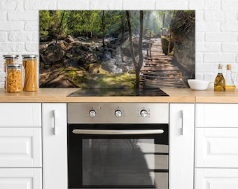 Puede incluir: Una cocina con armarios blancos y un horno de acero inoxidable. El protector contra salpicaduras es una imagen escénica de un puente de madera sobre un arroyo en un exuberante bosque verde.